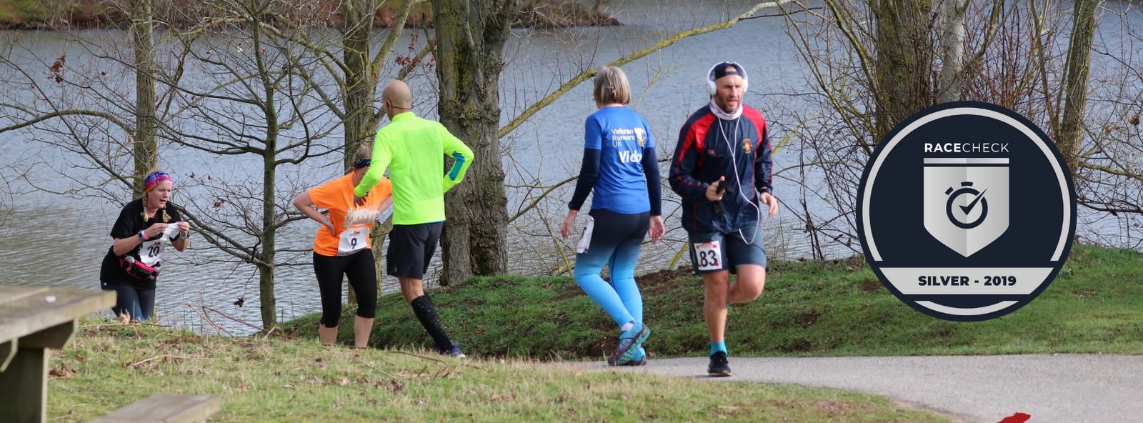 A diverse group of runners is participating in an outdoor race, passing by a lake with trees in the background. One runner, wearing a fluorescent yellow jacket, leads the group. A "Racecheck Silver - 2019" badge is shown on the right side of the image.
