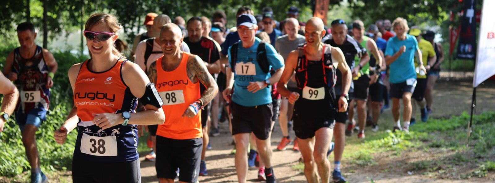A group of runners participating in a race on a sunny day, moving along a dirt path surrounded by greenery. The runners are wearing numbered bibs and various athletic gear, with one runner in front having the number 38.