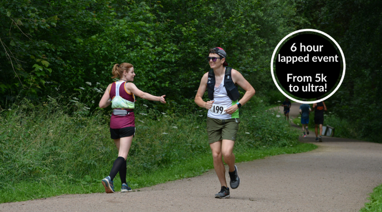 Two runners, a woman with a white headband and a man wearing a cap and sunglasses, share a high-five on a forested trail during a 6-hour race event. The man wears a bib numbered "199". A sign reads, "6 hour lapped event, From 5k to ultra!".