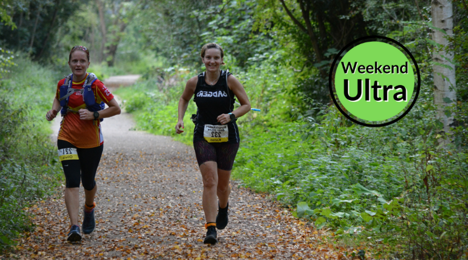Two women run on a forest trail during a race. One wears a red top with a backpack and the other wears a black top. Both have race bibs. The background is lush with greenery, creating a serene environment. A circular graphic with "Weekend Ultra" is in the top right corner.