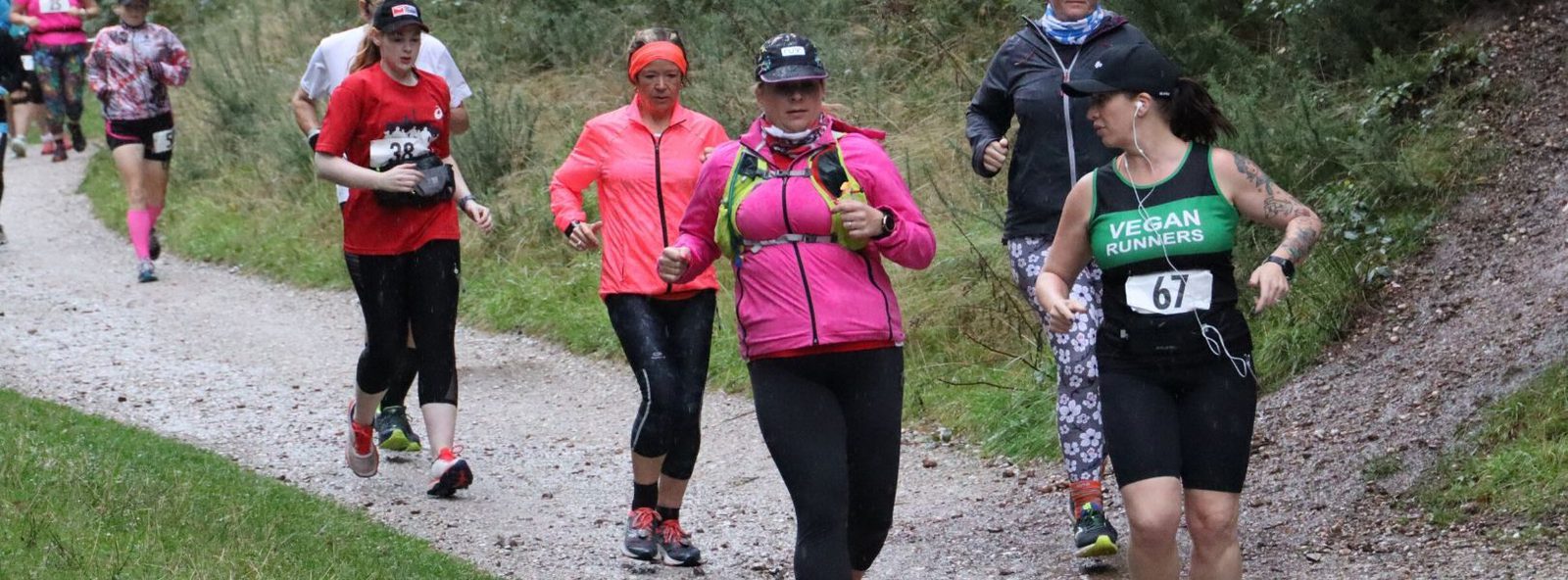 A group of runners on a trail in a forest setting. One leads wearing a green tank top and headband with "Vegan Runners" on the shirt, and a race bib numbered 67. Others wear varied outfits, including a red shirt, pink jacket, and black pants. The path is gravel, and the surroundings are green.