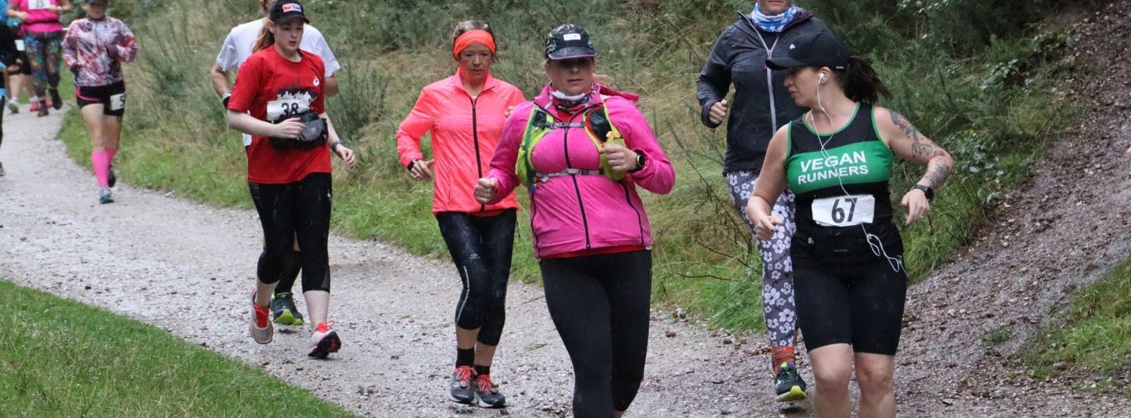 A group of runners on a trail in a forest setting. One leads wearing a green tank top and headband with "Vegan Runners" on the shirt, and a race bib numbered 67. Others wear varied outfits, including a red shirt, pink jacket, and black pants. The path is gravel, and the surroundings are green.