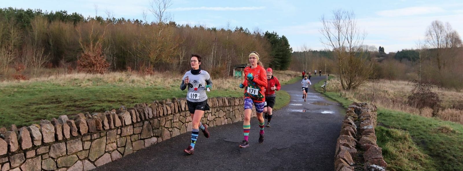 A group of runners is seen racing on a paved path surrounded by a natural landscape. The front runner wears a red shirt, followed by others in various athletic gear. The path is bordered by a stone wall on one side and grassy areas with trees and shrubs in the background.