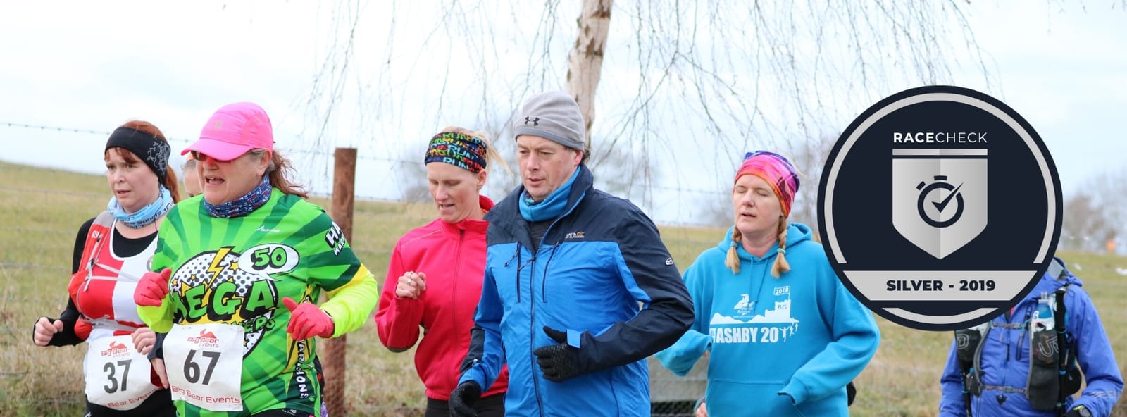 A group of runners, dressed in colorful athletic gear, are participating in a race. One runner wears bib number 67. The background shows a grassy field and bare trees. Overlaid on the right side of the image is a "Racecheck Silver - 2019" badge.