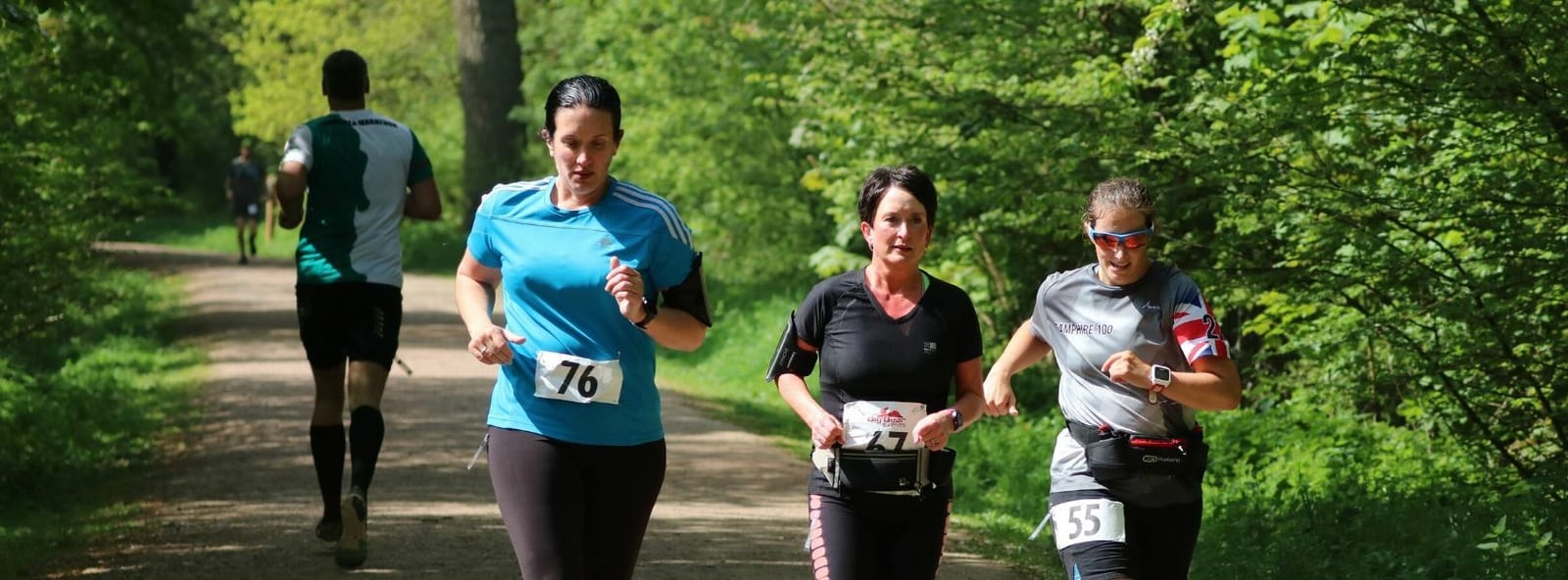 Three women are jogging together on a wooded trail during a race, wearing numbered racing bibs: 76, 5, and 55. They are focused and appear to be supporting each other. Lush greenery surrounds them, and a fourth runner is visible in the distance.