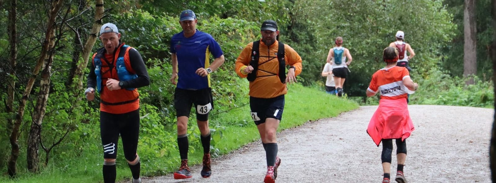 A group of runners competes in a trail race through a lush, green forest. Four runners in the forefront wear various colored shirts and hydration packs, and a few more are visible in the distance on the gravel path. The weather appears overcast and cool.
