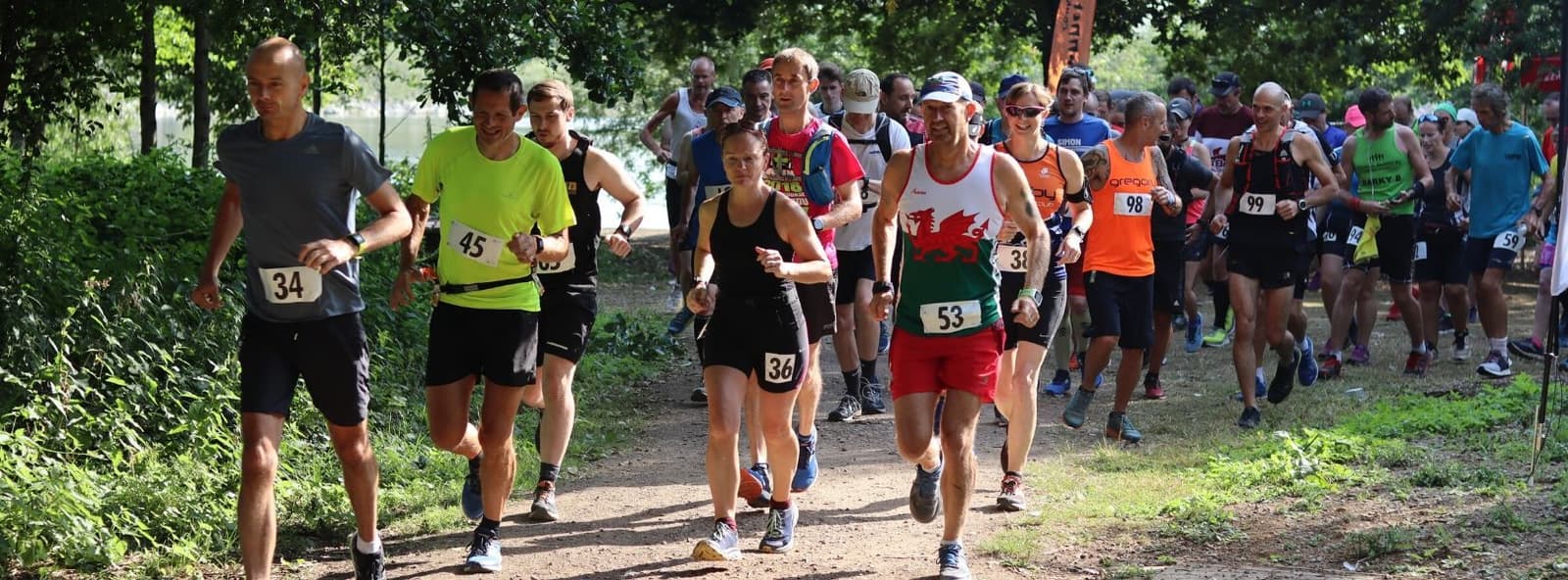 A group of runners, wearing numbered bibs, participating in a race on a forested trail. The runners are a mix of men and women of varying ages, dressed in athletic attire. The scene is daylight, and the path is surrounded by greenery and trees.