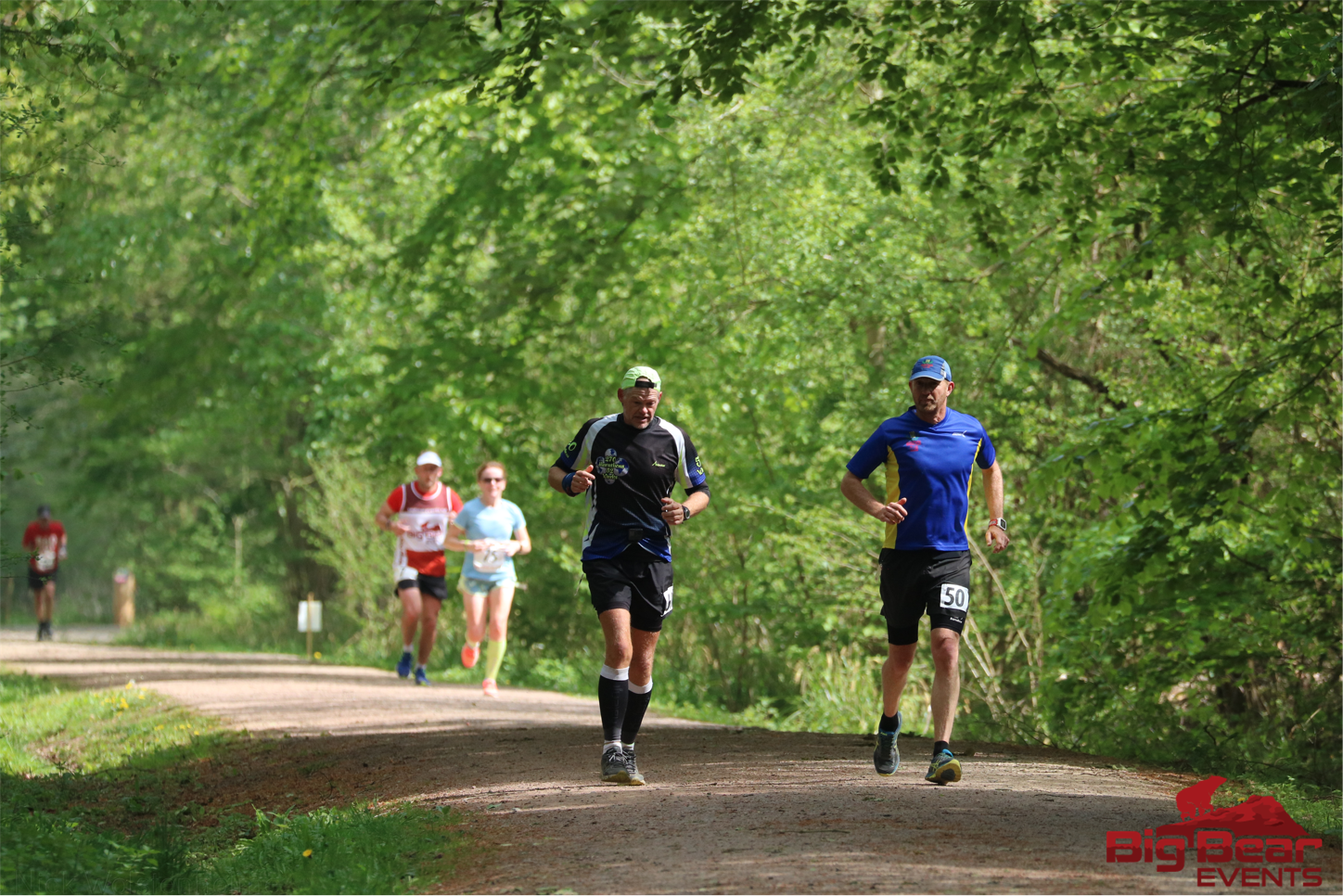 Five runners compete on a forest trail, surrounded by lush green trees. A man in a blue shirt and black shorts, wearing number 50, leads the group closely followed by another man in a black outfit. The event is organized by BigBear Events, as marked in the image.