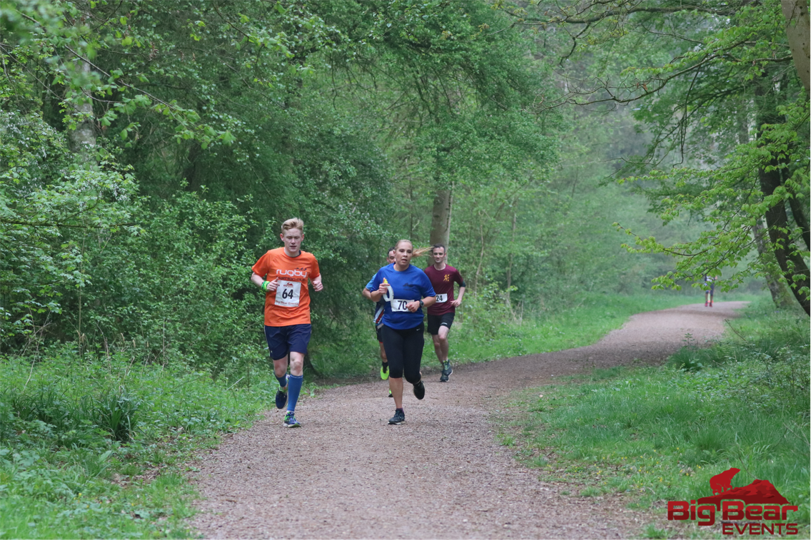 Three runners on a tree-lined trail during a race, two wearing blue shirts and one wearing a red shirt. They appear focused as they run, with lush greenery surrounding them. A fourth runner is seen in the distance. The event logo "BigBear Events" is visible in the bottom right.