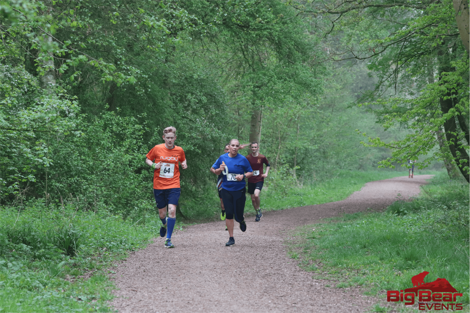 Three runners on a tree-lined trail during a race, two wearing blue shirts and one wearing a red shirt. They appear focused as they run, with lush greenery surrounding them. A fourth runner is seen in the distance. The event logo "BigBear Events" is visible in the bottom right.