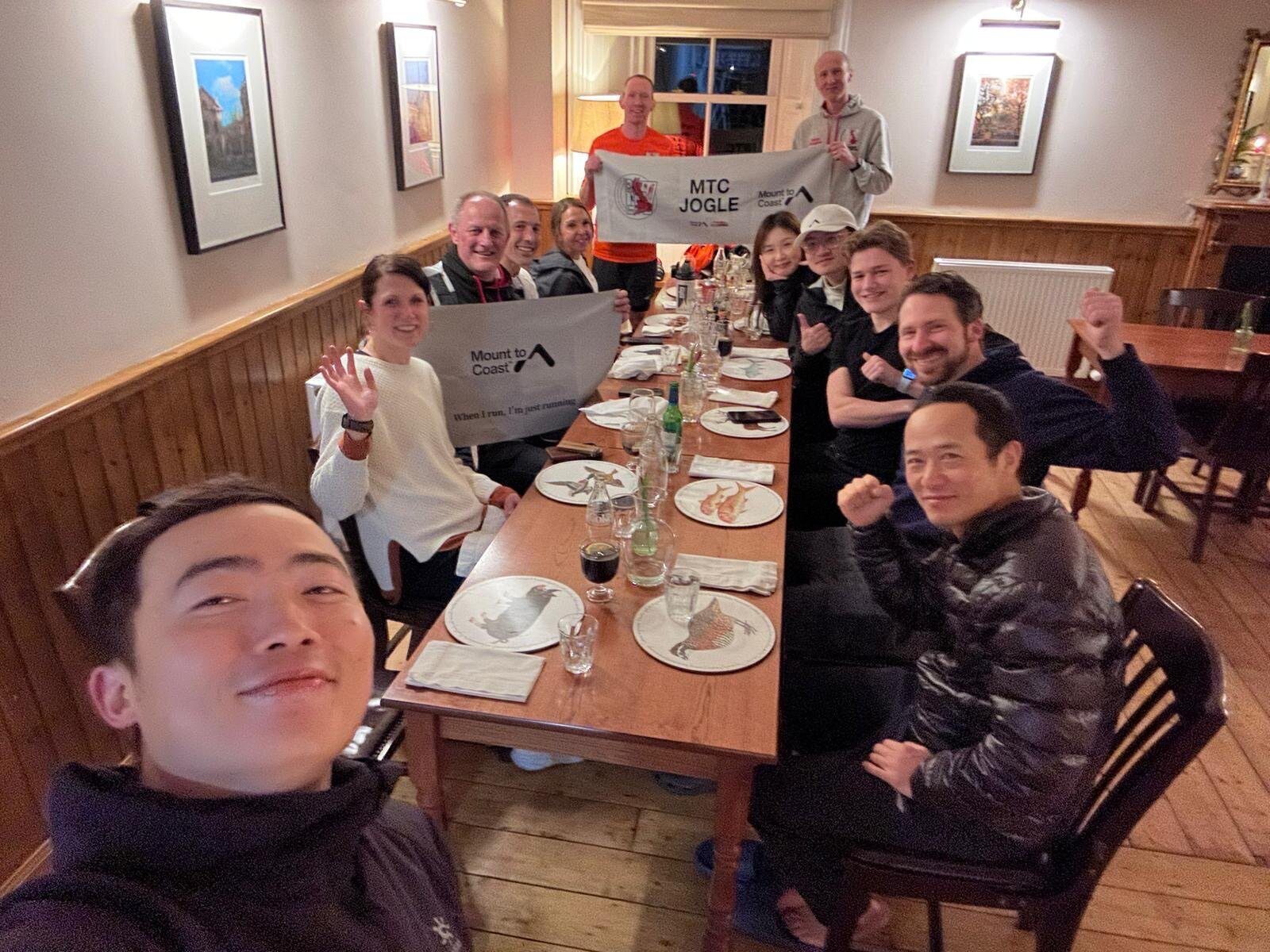 A group of people seated around a long wooden dining table in a cozy room, smiling and raising their fists in celebration. The table has plates and glasses. Two individuals at the back hold up banners, one reads "MTCJOGLE" and the other "Mount to Coast.