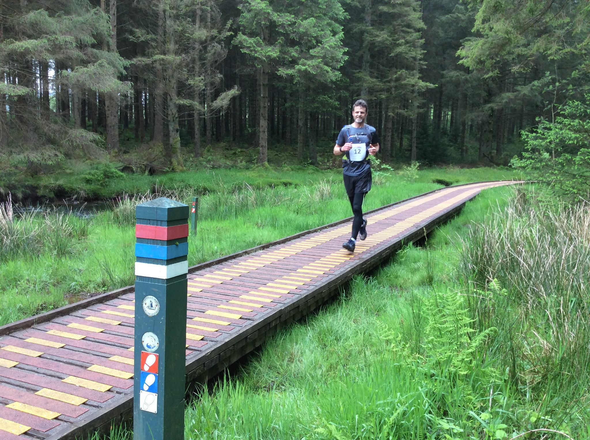 A man wearing a numbered bib jogs along a wooden path surrounded by lush greenery and dense forest. A colorful signpost with trail markers stands in the foreground, indicating a designated trail route through the woods.