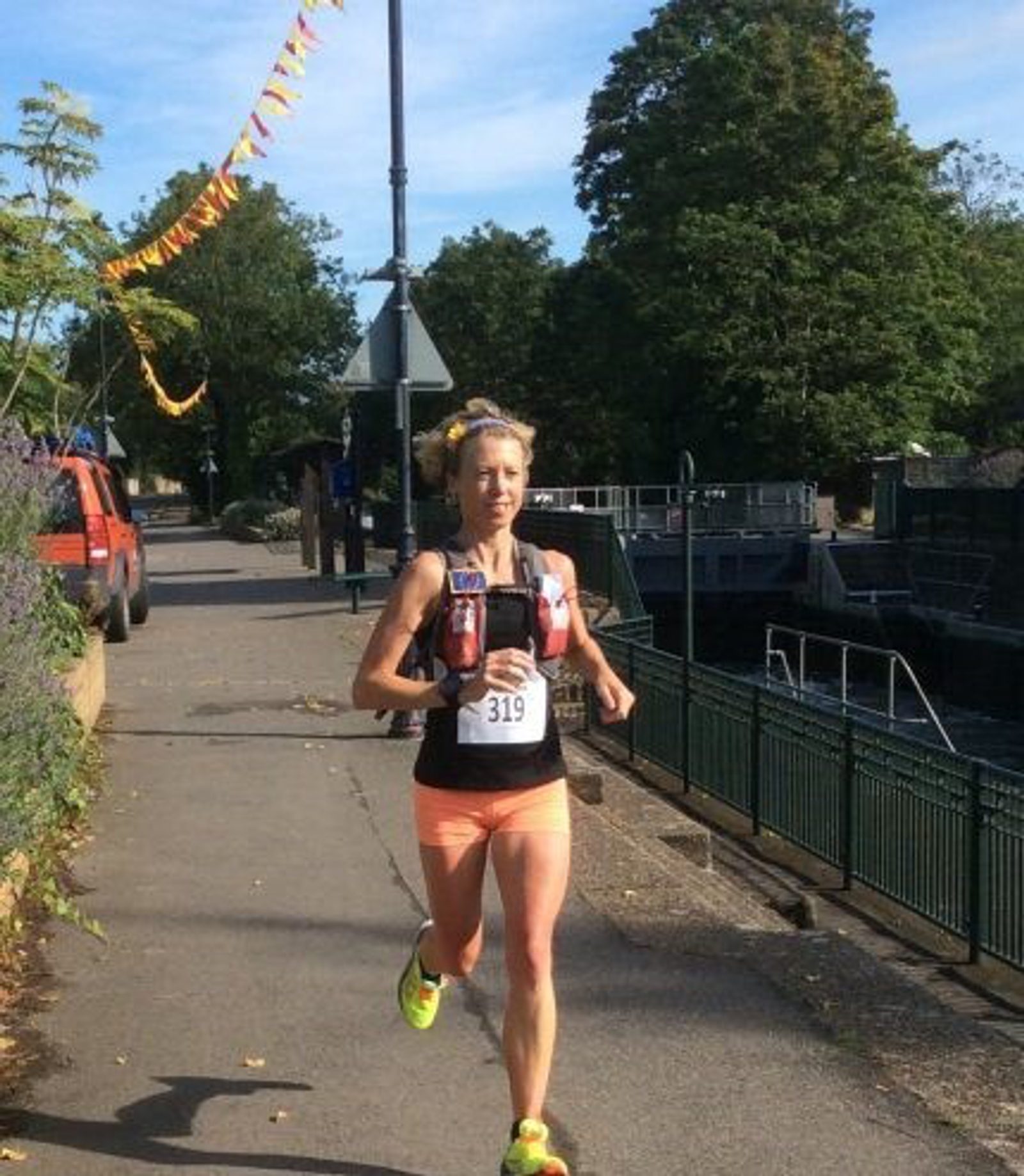 A person wearing a black tank top, orange shorts, and a race bib numbered 319 runs on a sidewalk in an outdoor setting with trees, a lamp post decorated with flags, and vehicles in the background. The individual also wears a hydration vest.