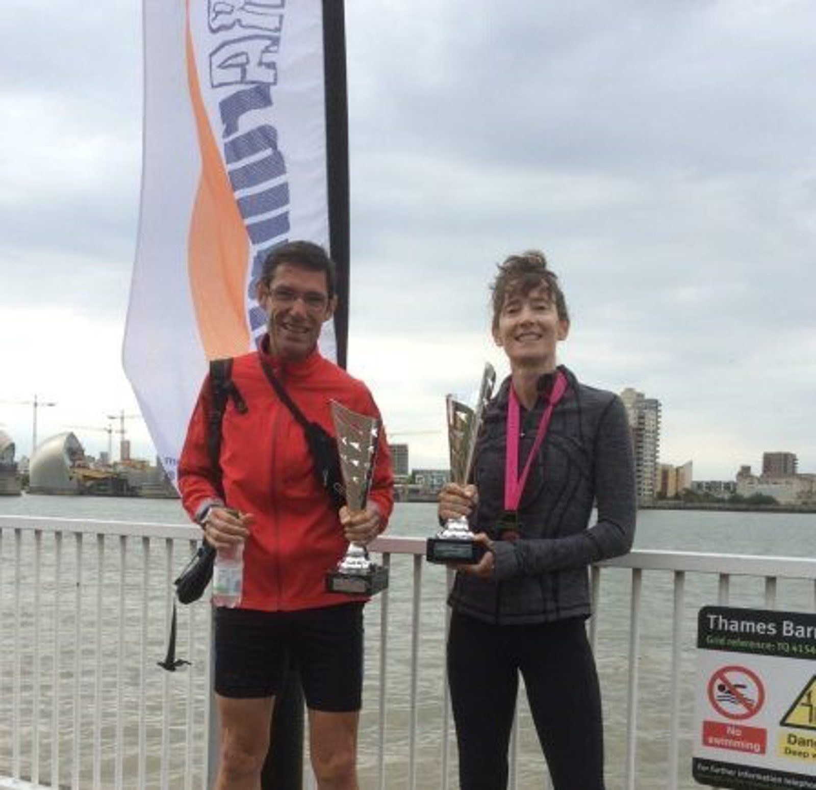Two people stand on a riverside, holding large trophies. Both are smiling and wearing athletic gear. A tall flag is visible in the background, along with buildings across the river. A sign with "Thames Barrier" is on the right side of the image.
