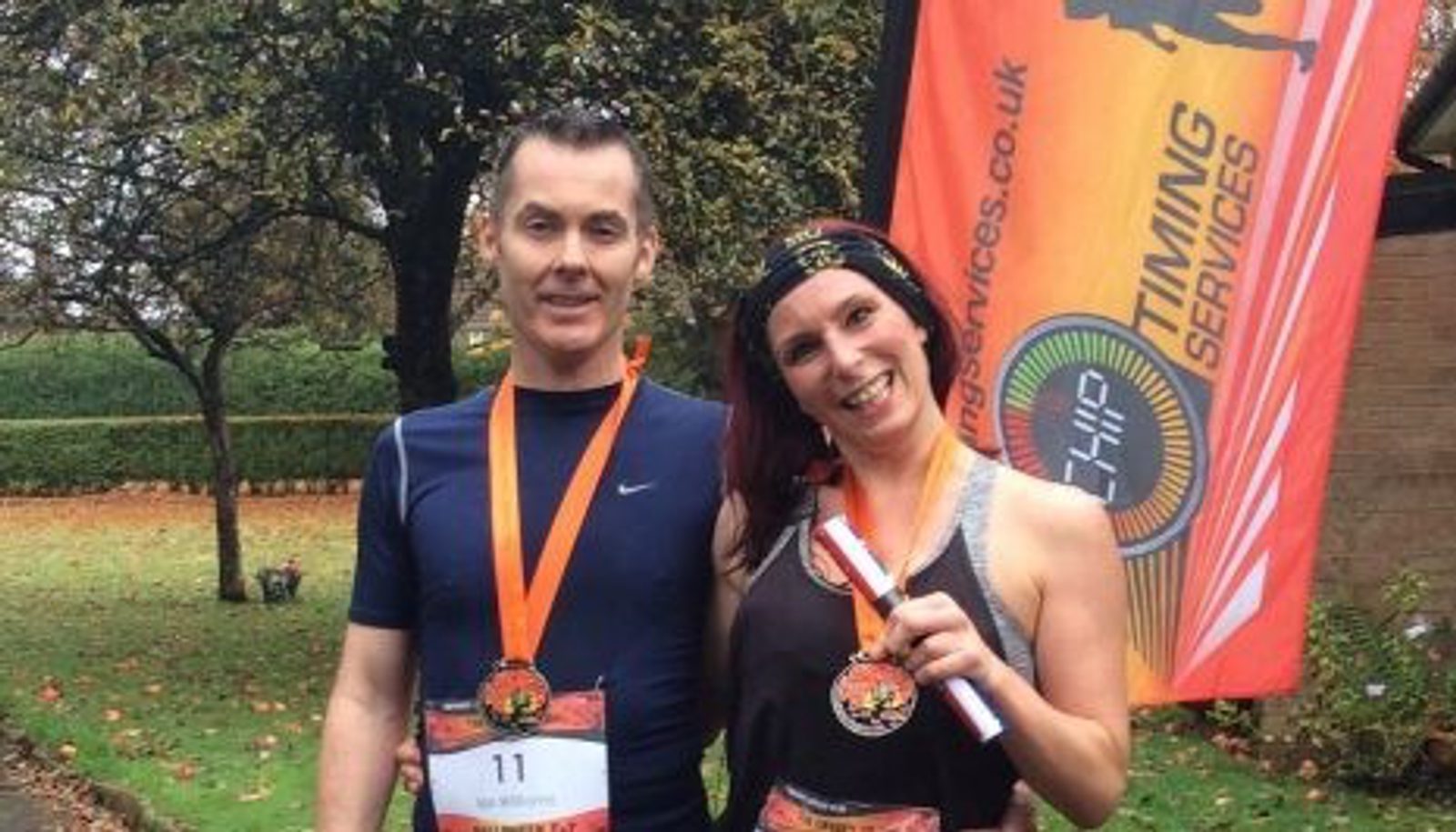 Two smiling individuals wearing orange race medals around their necks pose for a photo outdoors. The person on the left wears a black sleeveless top, and the person on the right, wearing a headband, holds up their medal. A bright orange banner is visible in the background.
