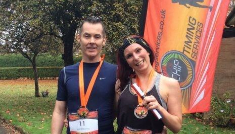 Two smiling individuals wearing orange race medals around their necks pose for a photo outdoors. The person on the left wears a black sleeveless top, and the person on the right, wearing a headband, holds up their medal. A bright orange banner is visible in the background.