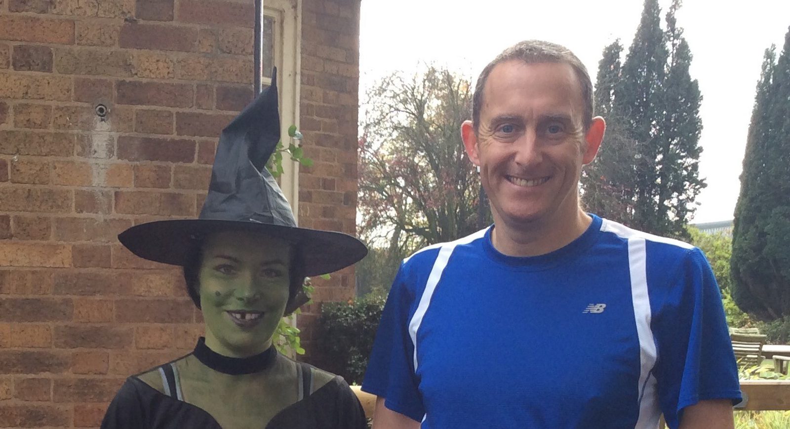 A smiling person in a blue athletic shirt stands next to another person dressed as a witch with green face paint, a pointy hat, and a black outfit. They are outside in front of a brick building with trees in the background.
