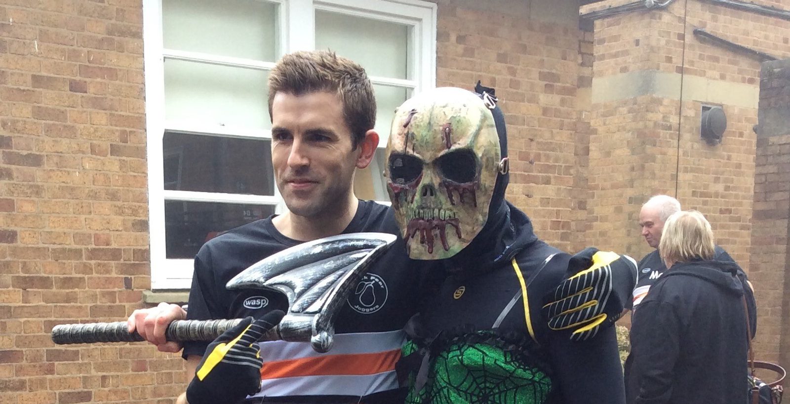 A man dressed in a black and white T-shirt poses with a Halloween-themed character wearing a grotesque skull mask and black outfit with green accents. The character holds a large silver weapon with a blade. They stand in front of a brick building.