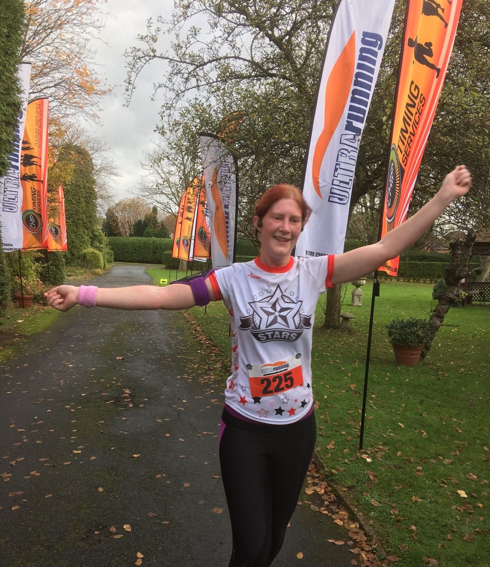 A person with light red hair, wearing a white shirt with a star design, black pants, and a race bib numbered 225, crosses a finish line with arms raised. Bright flags and trees line the road in the background, signaling a race event.