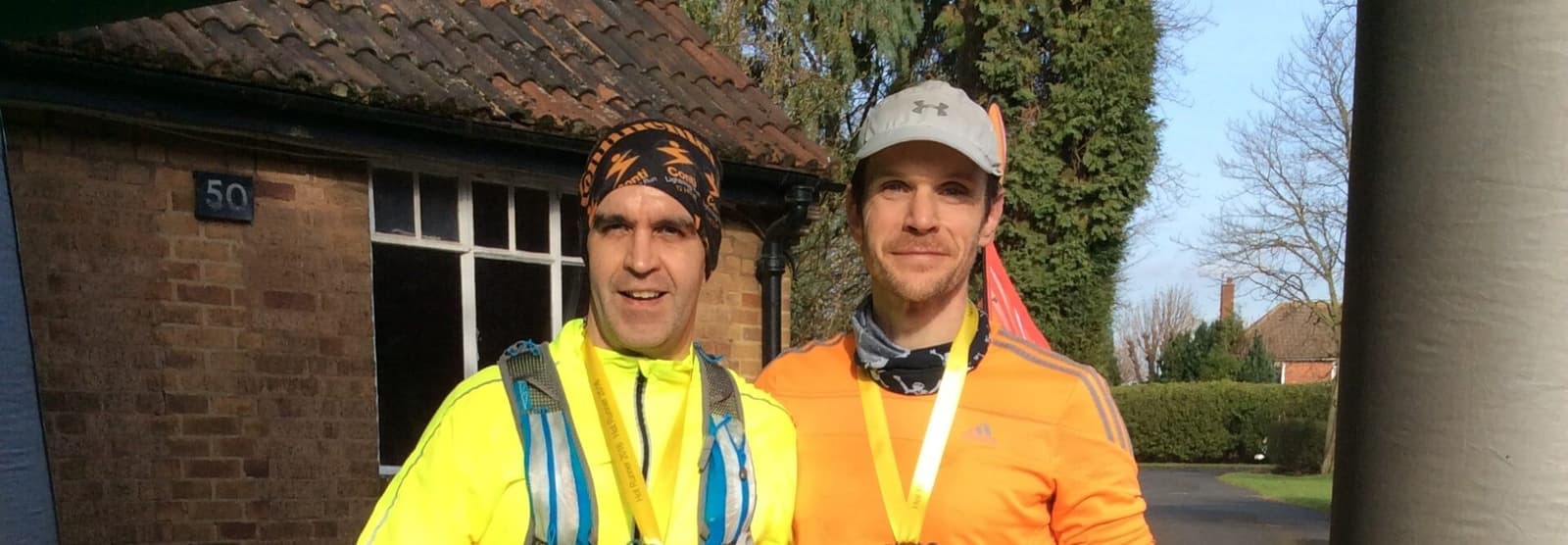 Two men stand side by side, each wearing a medal around their necks, likely after a race or sports event. They are dressed in bright athletic gear, with one in a yellow jacket and the other in an orange jacket. There is a building with a tiled roof and a tree in the background.