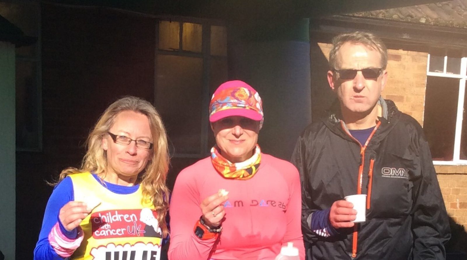 Three people are standing outdoors in bright sunlight in front of a building. Two women and one man are holding small cups. The woman on the left wears a "Children with Cancer UK" vest. The woman in the middle wears a colorful hat and pink top. The man wears sunglasses and a dark jacket.
