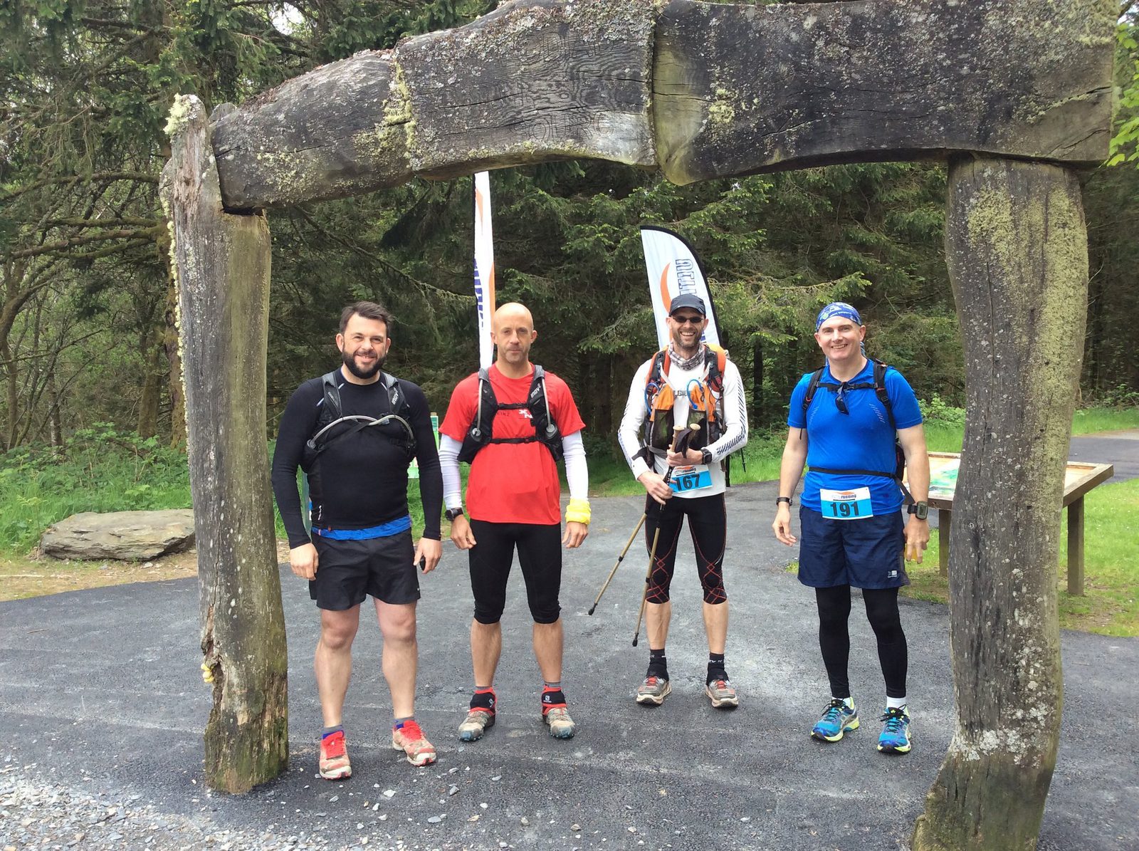 Four men in running gear stand under a rustic wooden archway in a forested area. They have race bibs on their shorts, and two of them are holding walking poles. The ground is paved, and there are trees and a flag banner in the background.