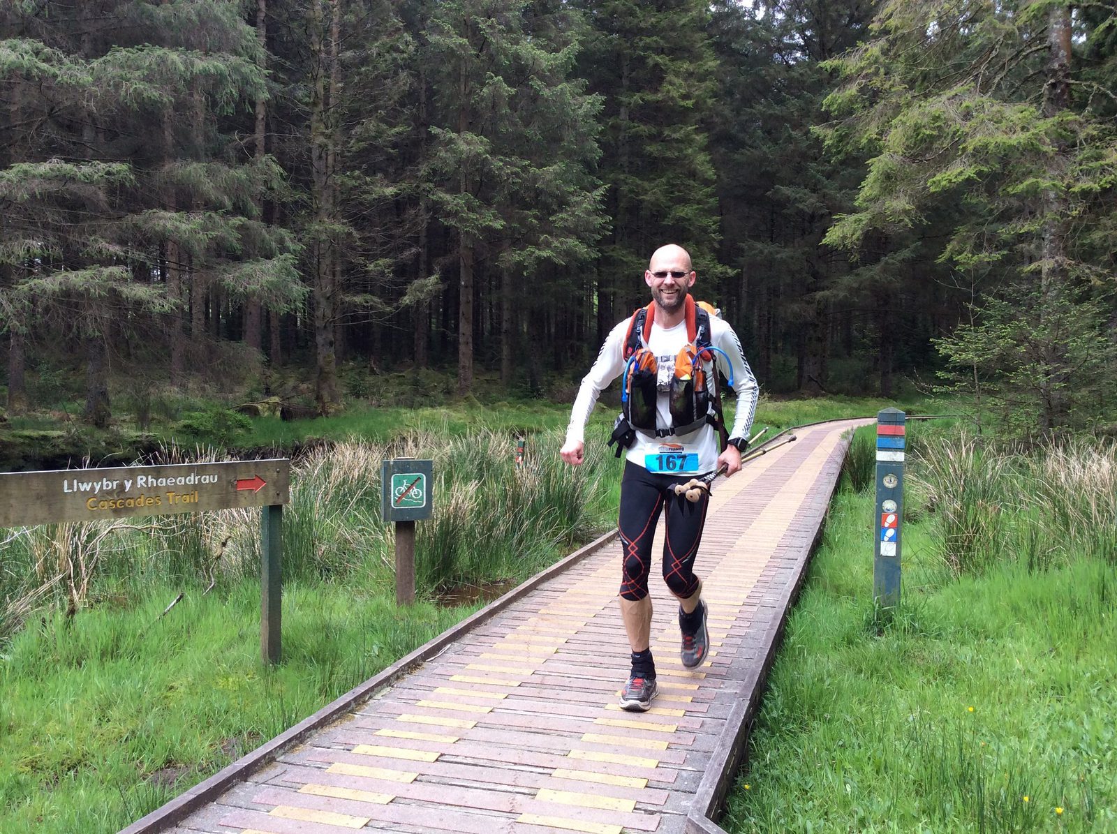 A man runs across a wooden bridge in a forest during a trail race. He wears a long-sleeve shirt, running tights, a hydration pack, and number 167 pinned to his shorts. The trail is called "Llwbyr y Rhaeadrau" according to the sign on the left.