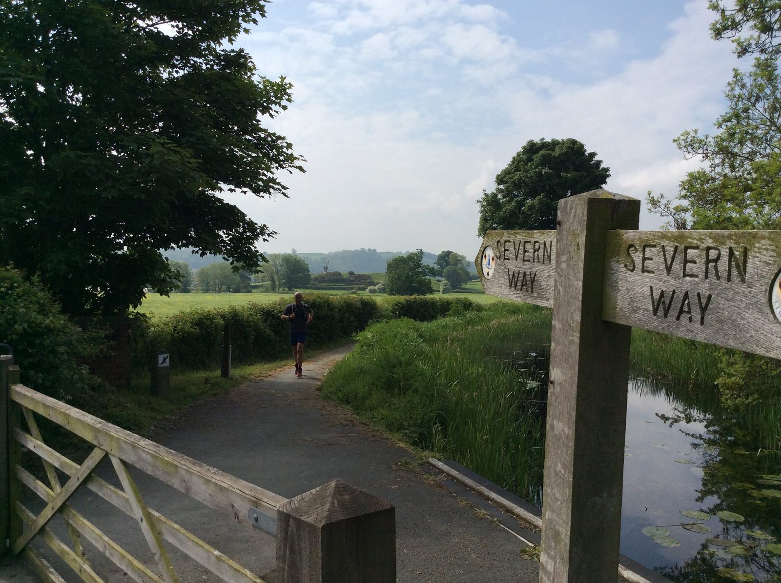 A person jogs along a paved path flanked by lush greenery and a wooden canal. Wooden signs on either side of the path read "Severn Way." Trees and open fields are visible in the background under a partly cloudy sky.