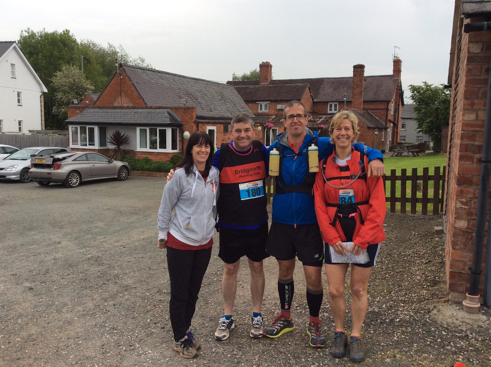 Four people are standing together outdoors, smiling at the camera. They are dressed in sportswear and running gear, with two of them wearing race bibs labeled 180 and 44. The background features a few brick buildings and a parked car. It looks cloudy.