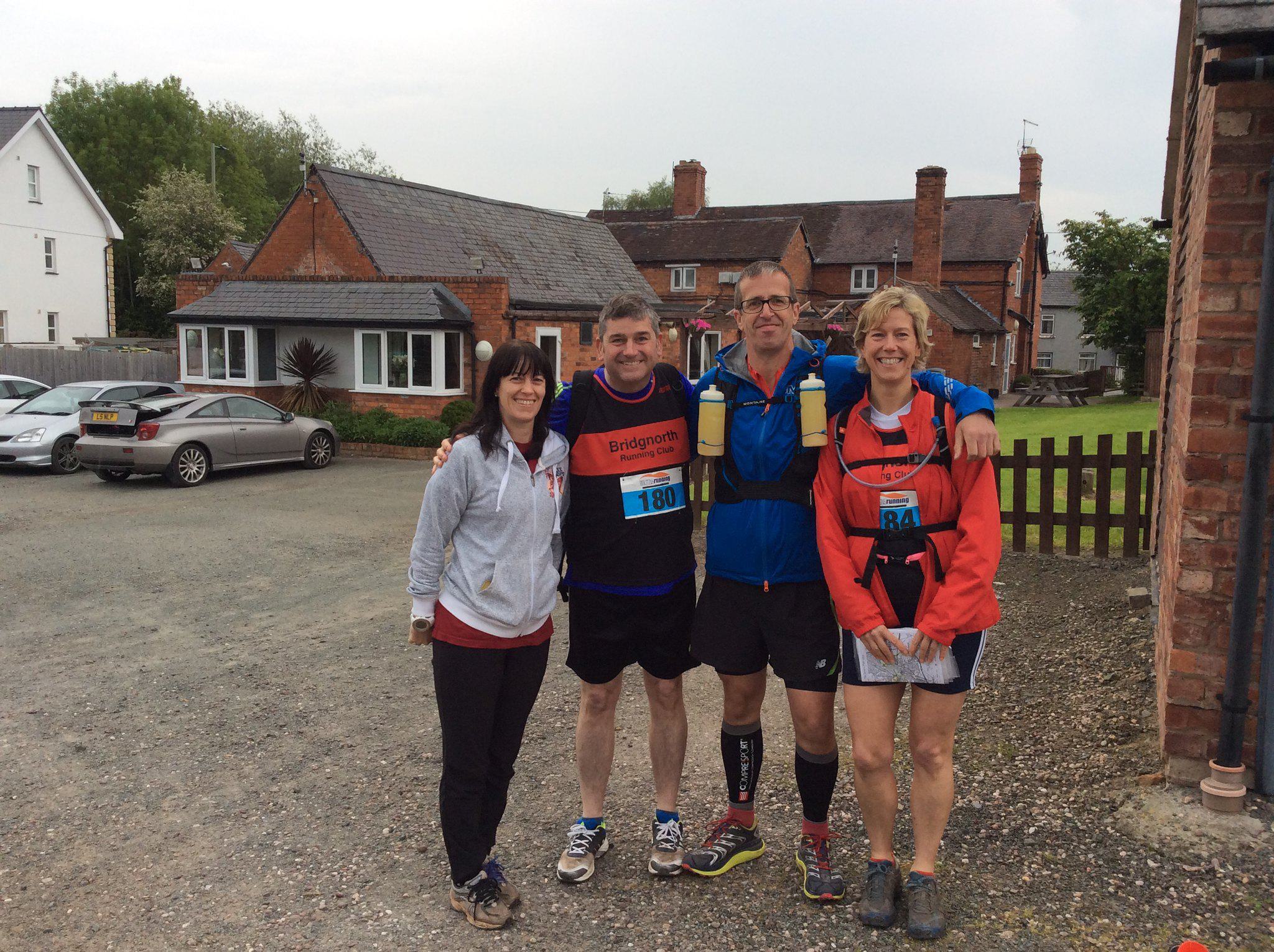 Four people are standing together outdoors, smiling at the camera. They are dressed in sportswear and running gear, with two of them wearing race bibs labeled 180 and 44. The background features a few brick buildings and a parked car. It looks cloudy.