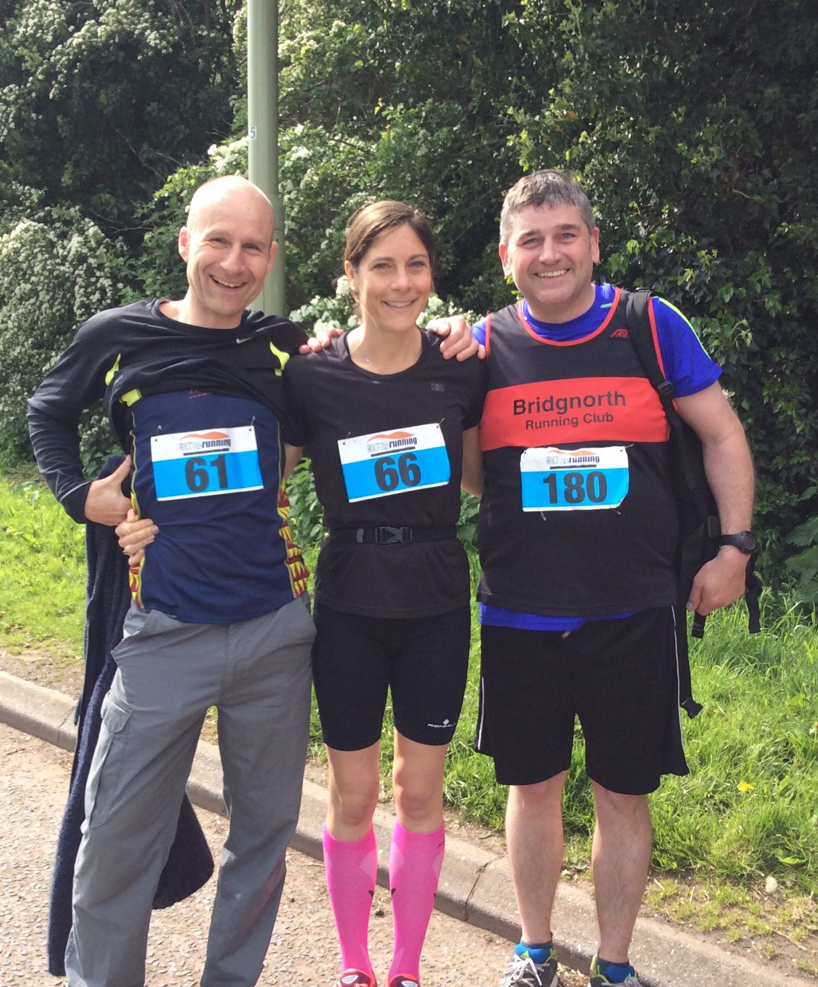 Three runners, two men and one woman, posing together after a race. Each wears a race bib with numbers 61, 66, and 180 respectively. The woman is in the center, wearing bright pink socks, and the man on the right is wearing a Bridgnorth Running Club shirt. They are smiling.
