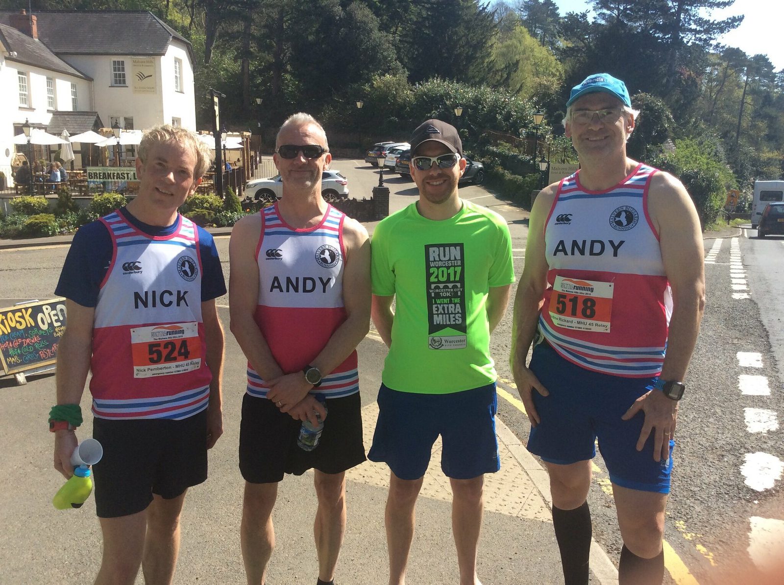 Four runners are posing for a photo on a sunny day. Three wear red and white striped club vests with "ANDY" and "NICK," along with race bibs. The fourth wears a green "RUN 2017" shirt and sunglasses. They are standing near a street with buildings and trees in the background.