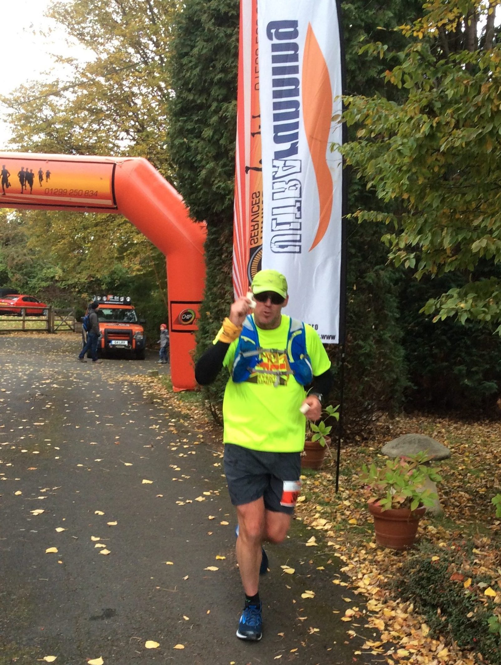 A runner, wearing a fluorescent yellow shirt and neon green cap, jogs past an orange inflatable archway near a "Ultra Running" banner. The scene is outdoors, along a tree-lined path with autumn leaves scattered on the ground.