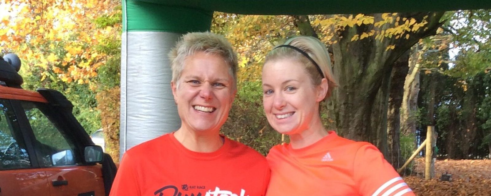 Two women in athletic attire stand smiling under a green and white canopy. The woman on the left has short blonde hair and wears a red shirt, while the woman on the right has long blonde hair and wears an orange shirt and a headband. Autumn trees are in the background.