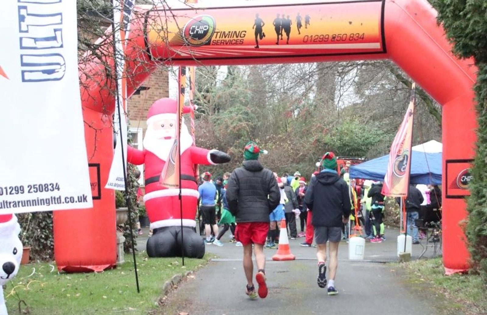 Two runners approach the starting arch of a race, which is decorated with a large inflatable Santa Claus and festive banners. Many participants in colorful athletic gear are gathered at the back. Trees and a building are visible in the background.