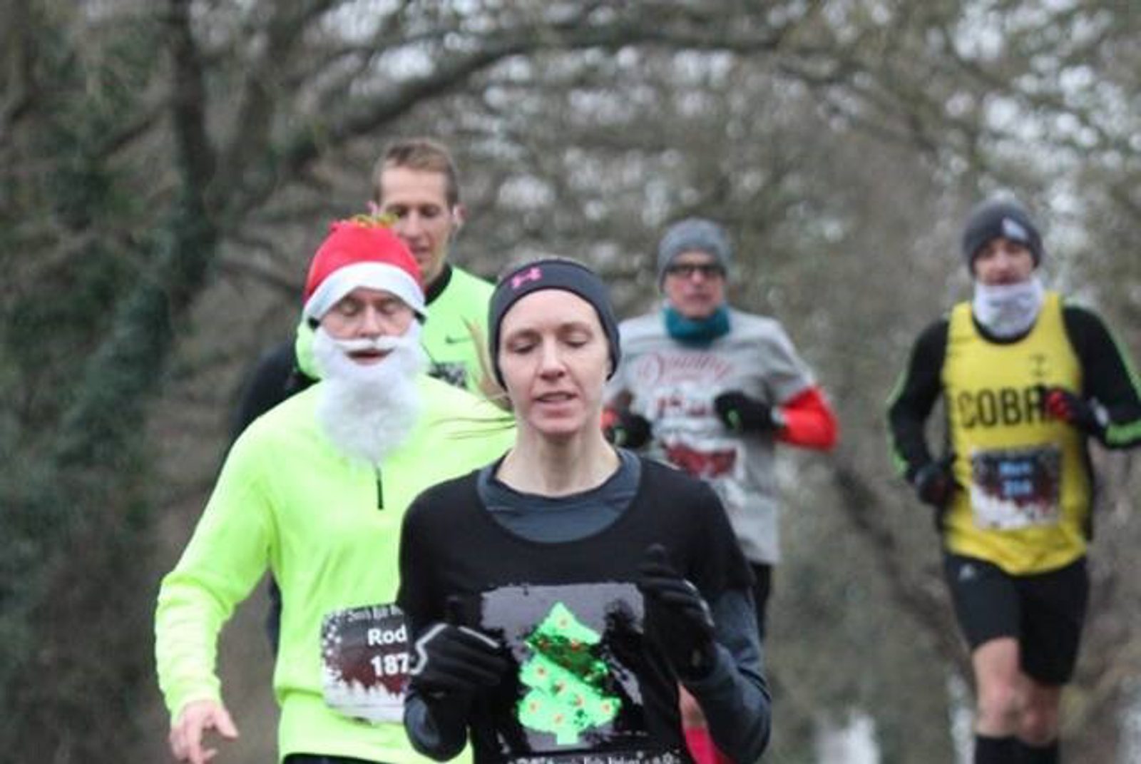 A group of people running outdoors during a winter race. One runner, wearing a neon green shirt and a Santa hat with a white beard, stands out. Another runner in the foreground is wearing a black shirt with a Christmas tree on it. Trees are in the background.