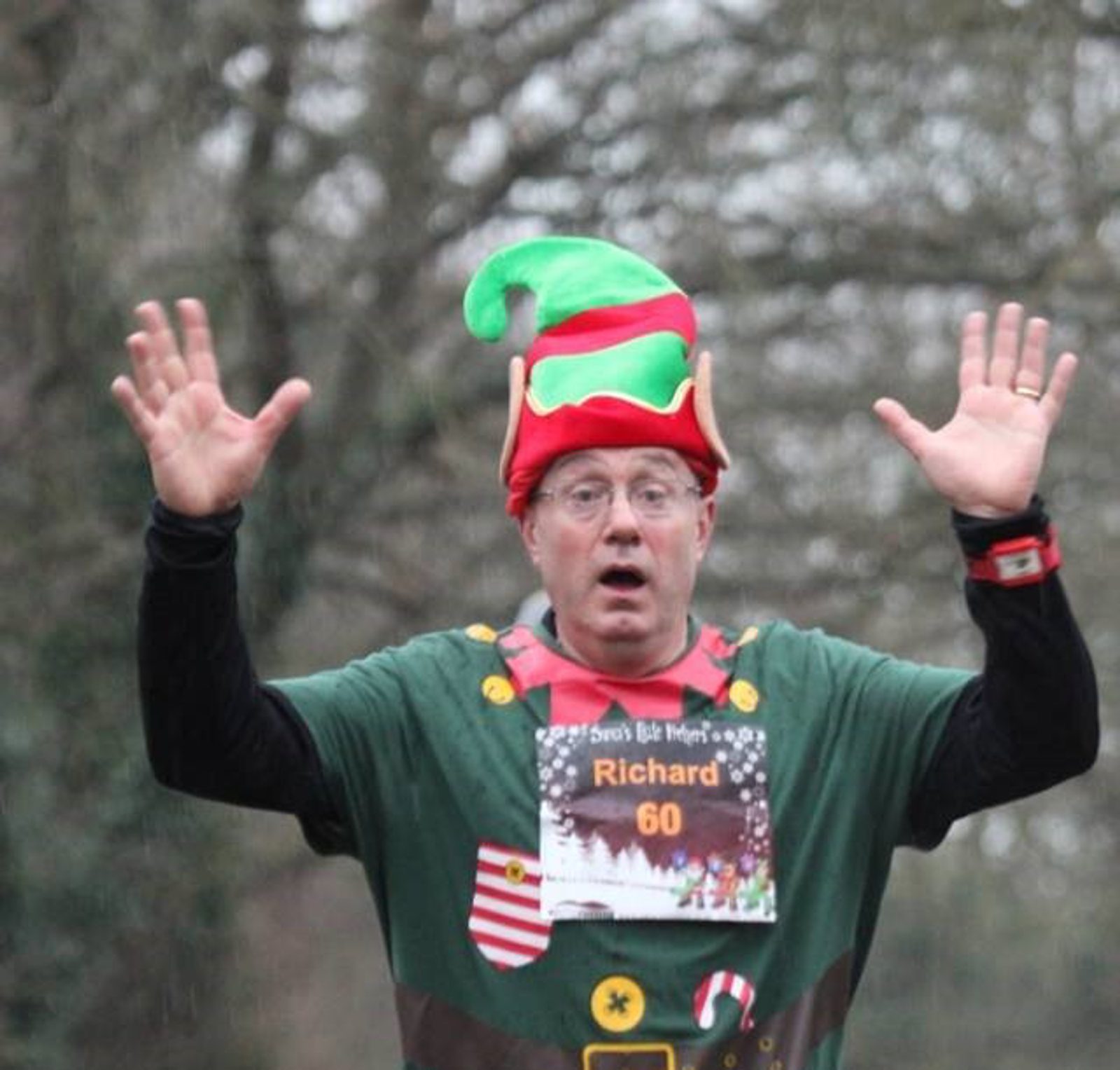 A man dressed in a festive elf shirt and hat is running outdoors with both hands raised. He has a race bib that reads "Richard 60" pinned to his shirt. The background features trees and greenery.