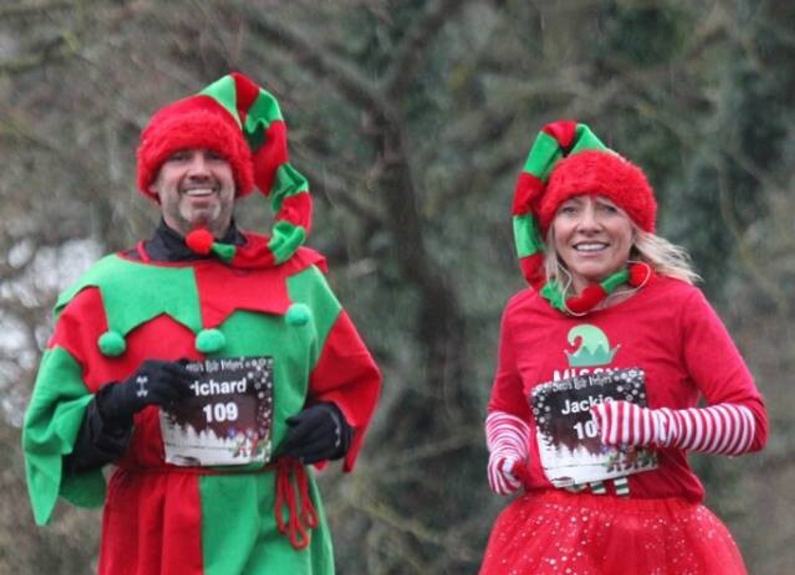 Two people, dressed as festive elves, run outdoors. Both wear red and green clothing with elf hats, a bib labeled "Jachard 109," and holiday-themed accessories like striped sleeves and tutu. They are smiling and appear to be participating in a holiday-themed race or event.