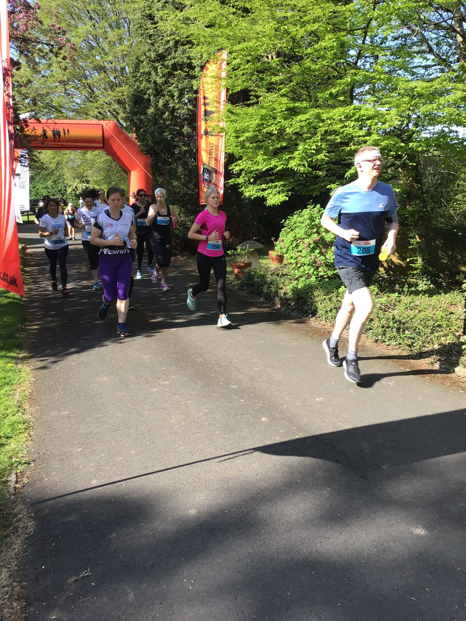 Runners participate in a race on a paved path surrounded by greenery. An orange inflatable arch marks part of the course. The scene is brightly lit by sunlight, and participants wear a variety of athletic clothing and bib numbers.