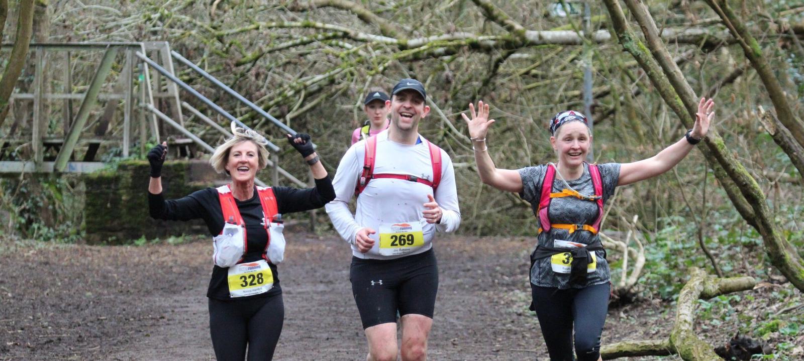 Three runners are jogging on a forest trail, smiling and raising their arms in celebration. They are wearing numbered race bibs and hydration packs. A wooden bridge and leafless trees can be seen in the background.