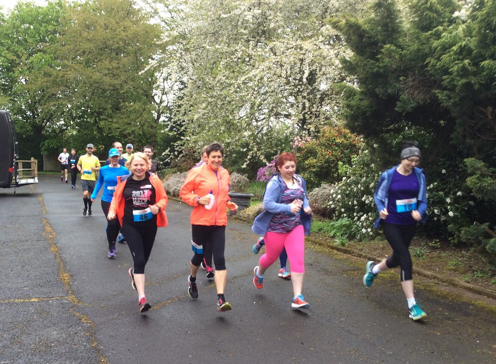 A group of runners, dressed in various athletic gear, jog along a paved path lined with trees and bushes. Some trees are blooming with white flowers. The weather appears cool, as some runners wear jackets and hats. Everyone looks focused and determined.