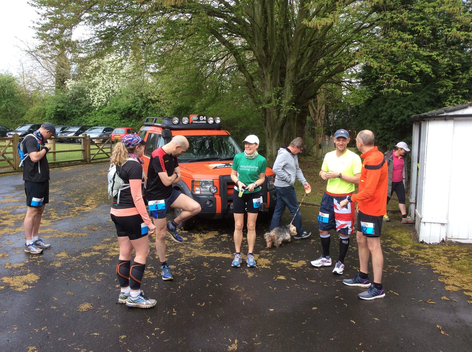 A group of runners wearing athletic gear gather near an orange vehicle and some trees. They appear to be preparing for or finishing a run, and a small dog is nearby. The ground is wet and covered with some fallen leaves. Some runners have race bibs attached.