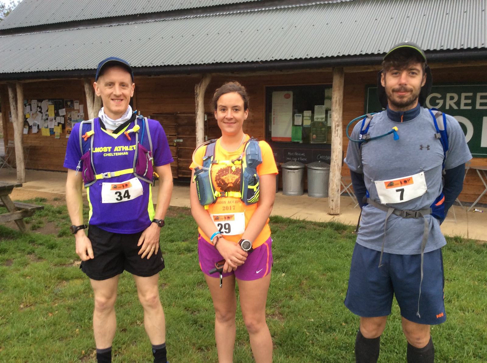 Three runners are standing in front of a wooden building. The runner on the left is wearing a purple shirt with the number 34, the runner in the middle is wearing a pink outfit with the number 29, and the runner on the right is in a blue outfit with the number 7.