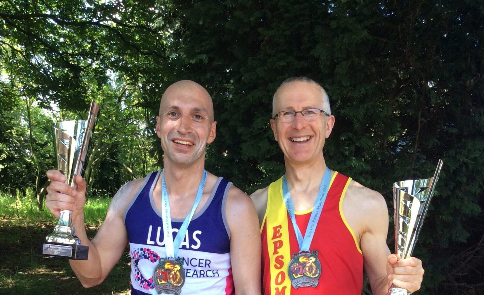 Two smiling men hold trophies and display their medals while wearing running gear. The man on the left has a shirt that reads "CANCER RESEARCH" with the name "LUCAS" above it. The man on the right is wearing a shirt with "EPSOM" written on it. Trees are in the background.