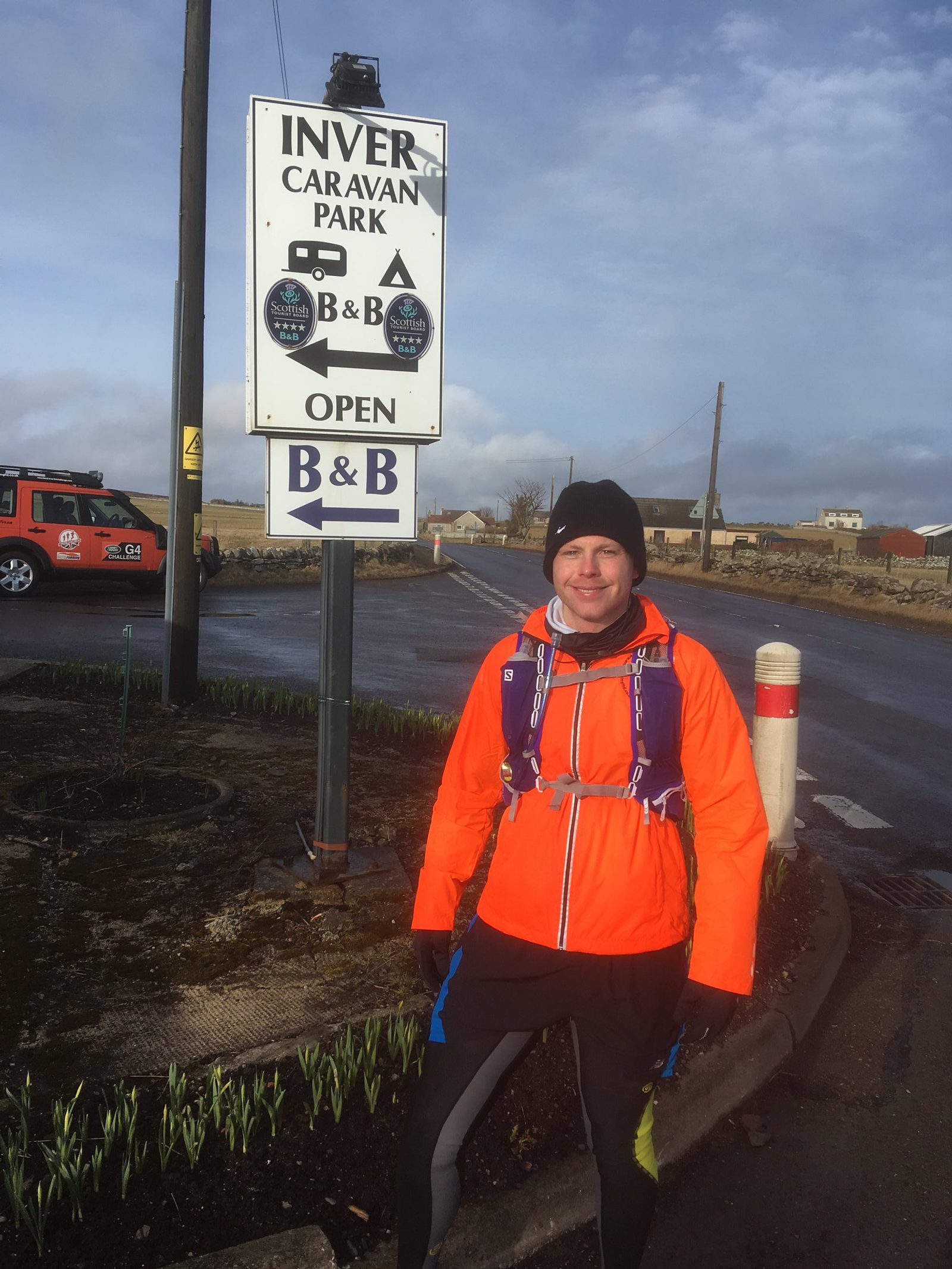 A person wearing a bright orange jacket, black pants, and a black beanie stands on a paved area near an "Inver Caravan Park" sign. The sign indicates the availability of B&B services. In the background, there is a red vehicle and a rural landscape.