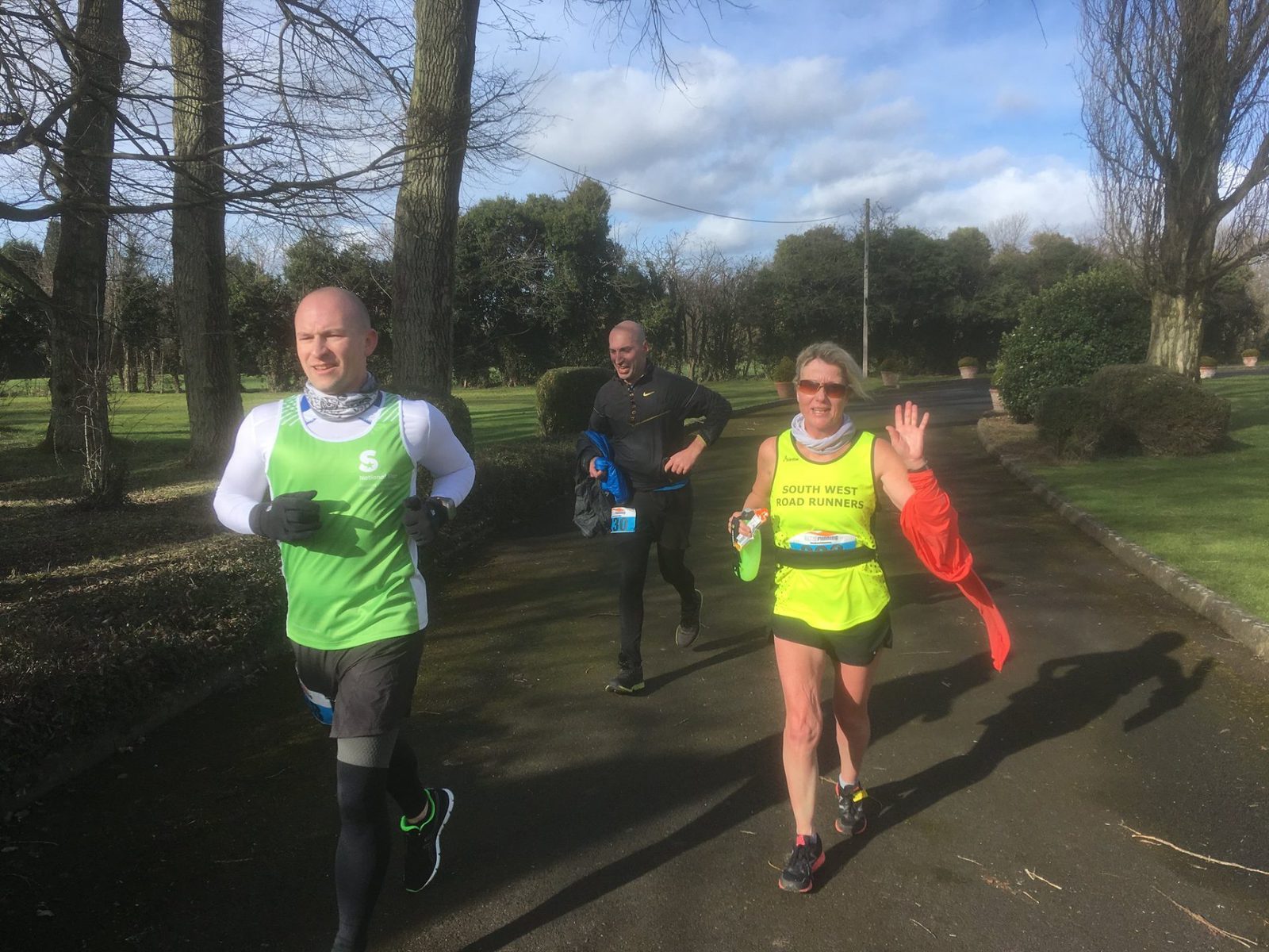 Three runners on a paved path in a park, with trees and bushes around them. The runner on the right is waving and smiling, wearing a yellow and green outfit. The middle runner is holding a water bottle, and the left runner is wearing a green vest and black leggings.