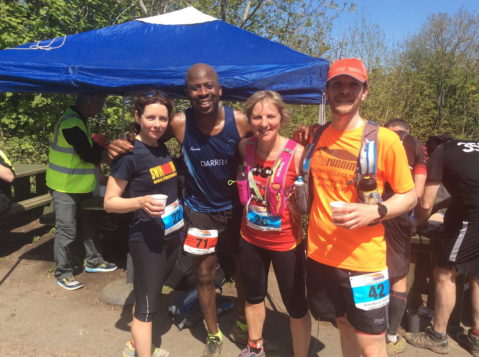 Four runners stand in front of a blue canopy, smiling at the camera. The man in the middle is wearing running gear that says "Darren." Two women stand on either side of him, and another man is on the far right. They are holding water cups, likely during a race or marathon.