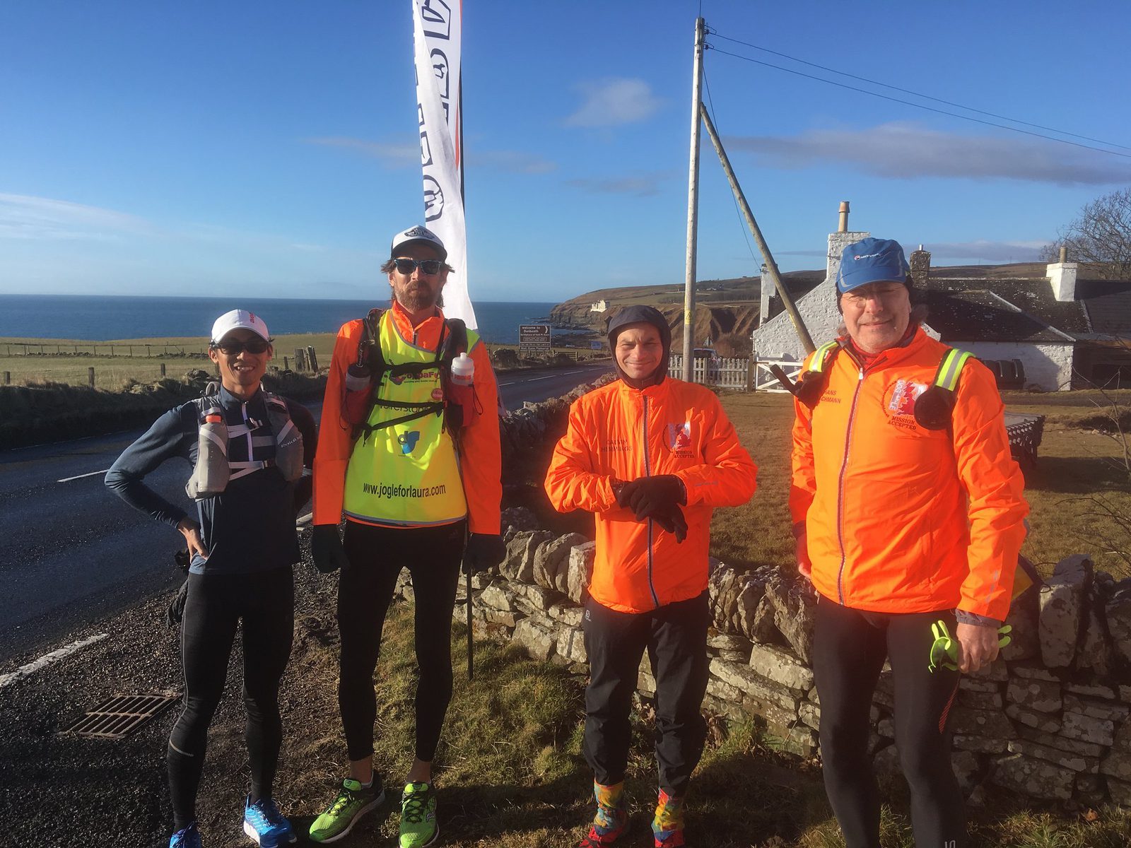 Four runners posing outside next to a stone wall. They are wearing brightly colored running gear and backpacks. A flag with text and logos is behind them. In the background, there's a road, grass, houses, and a view of the sea under a sunny blue sky.