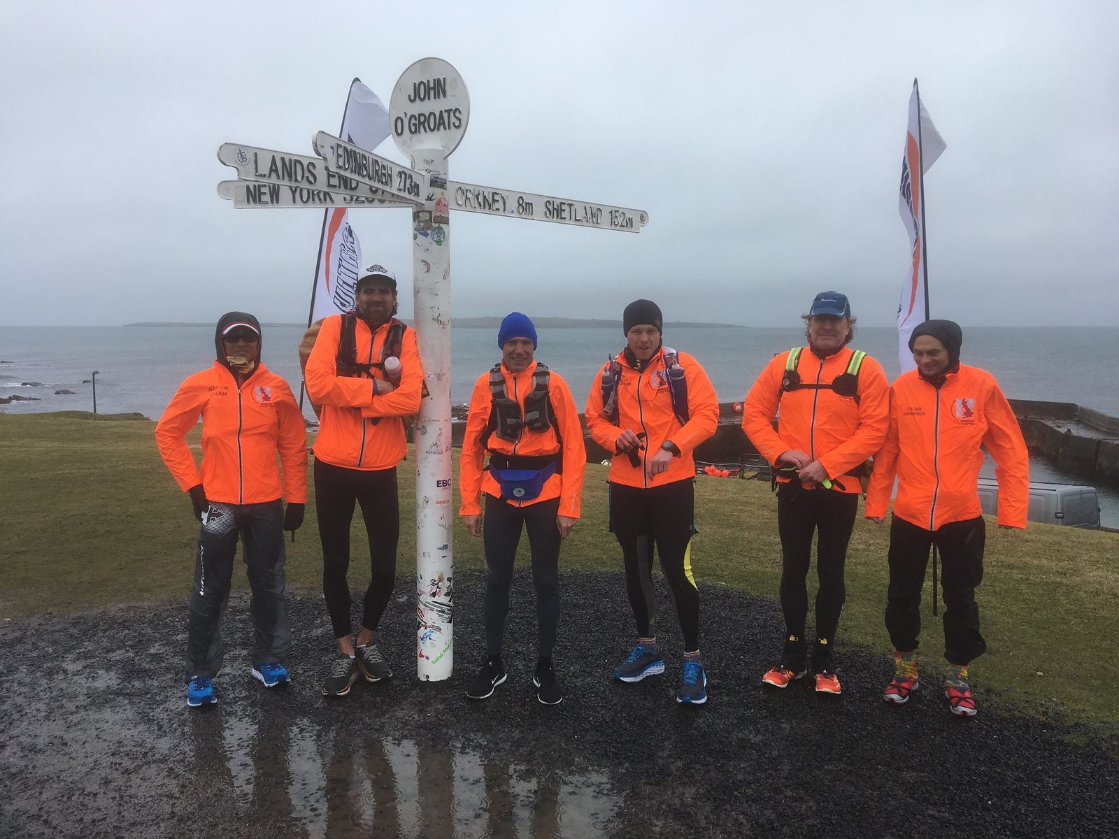 Six individuals wearing bright orange jackets with backpacks stand in front of a white signpost at John O'Groats. The overcast sky and ocean in the background create a moody atmosphere. The signpost indicates various distances to notable locations.
