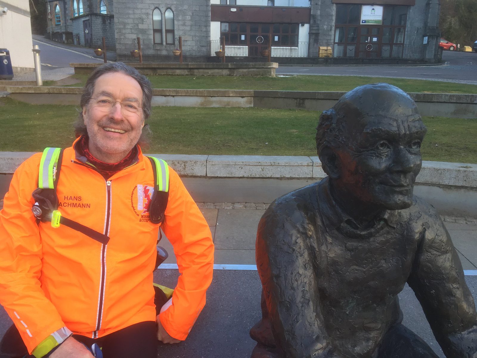 A man with long hair and glasses, wearing an orange jacket, sits next to a bronze statue of an elderly man. They are outside in front of a building with stone walls and arched windows. The man is smiling at the camera, and the statue has a neutral expression.